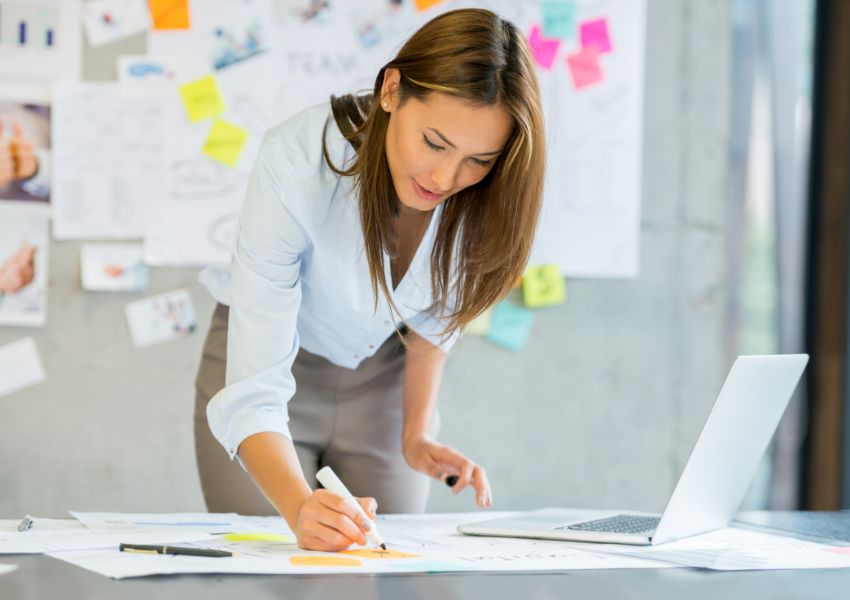 A landlord with long brown hair stands over a desk as they make their business plan using paper, a laptop and post its.